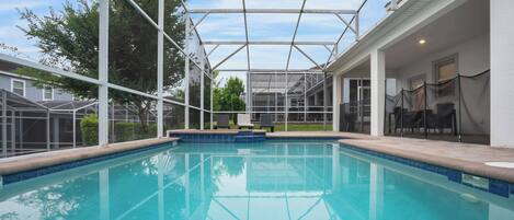 A swimming pool with a covered area, surrounded by a white building with a patio and outdoor seating.