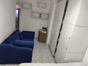 A room with a blue sofa set, a wooden side table, a white wardrobe with gold trim, and a small shelf with decorative items. - Cozy apartment in a great location. (Praia Grande)