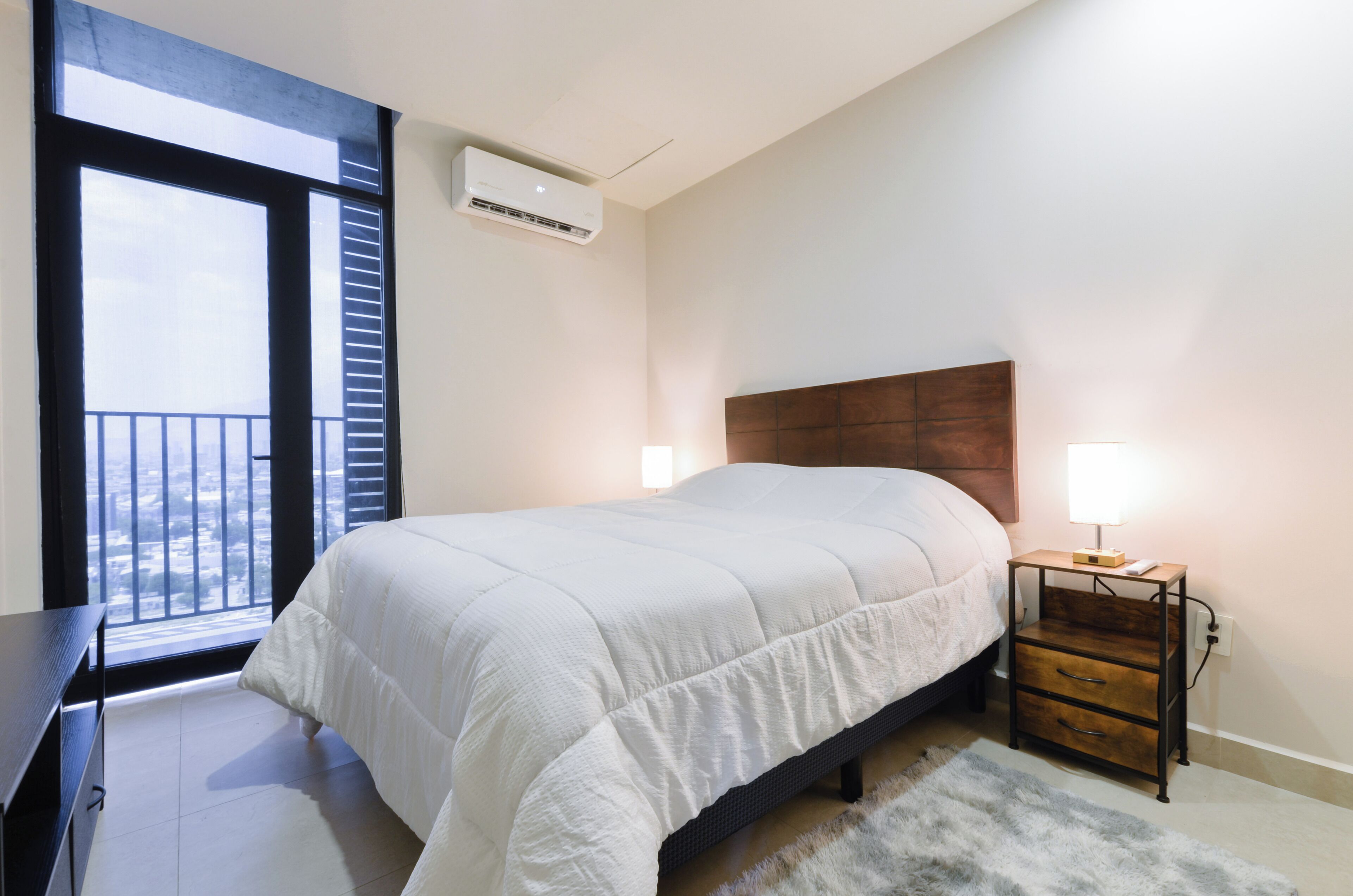 A bedroom with a bed, bedside tables, a nightstand, a window with blinds, and an air conditioning unit.