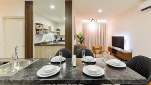 Comedor con una mesa de mármol preparada para cuatro personas, una cocina al fondo y una sala de estar con televisor y sofá.