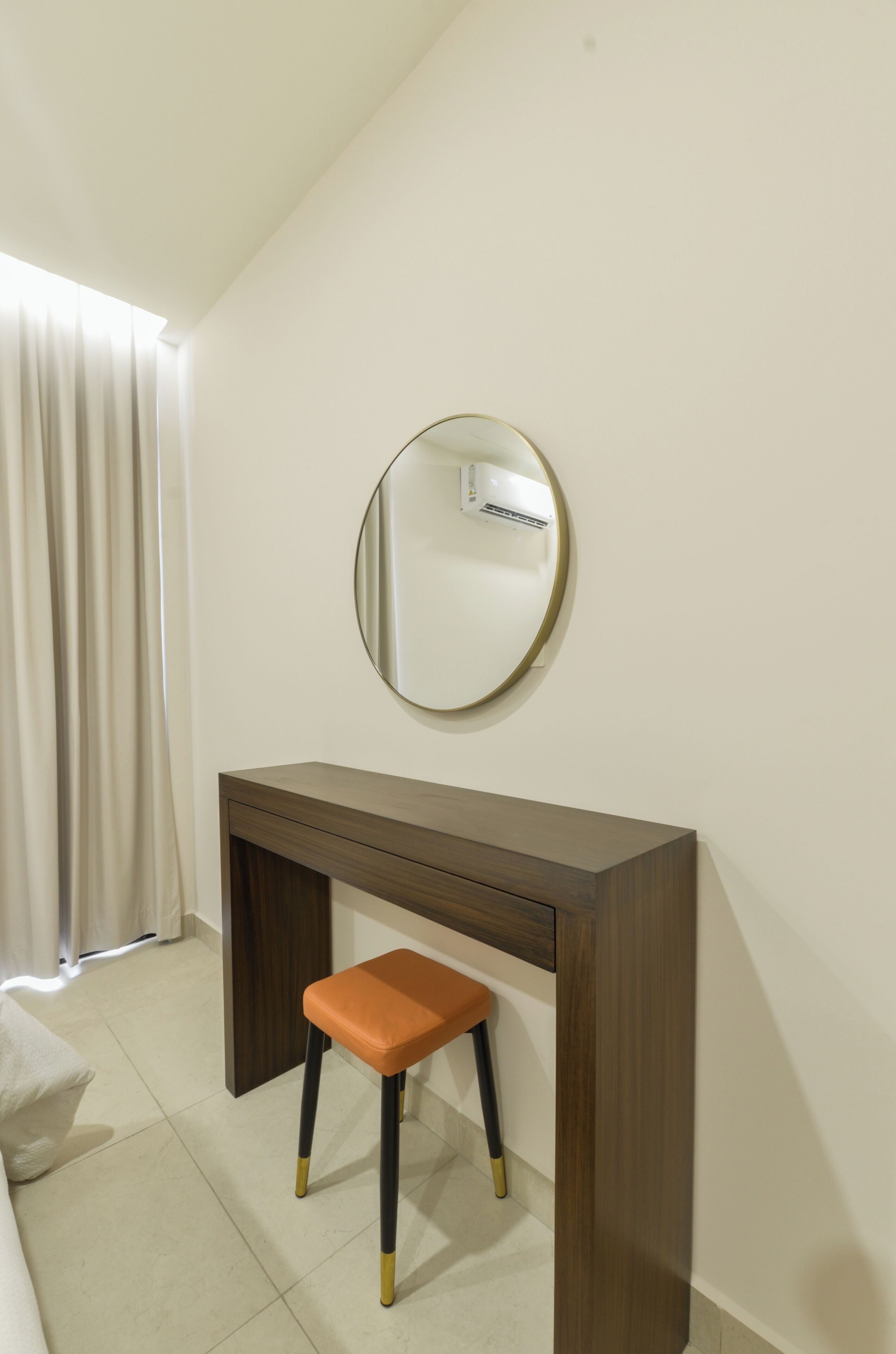 Una habitación con escritorio, taburete, espejo y ventana con cortinas.