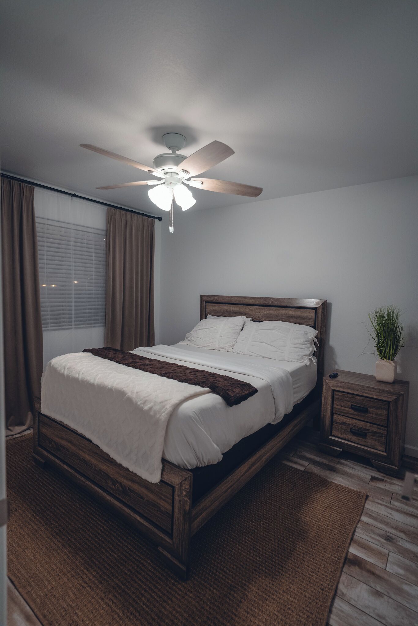 A bedroom with a bed, a ceiling fan, a bedside table with a plant, and a window with curtains.