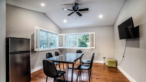 Une pièce comprenant une table à manger, des chaises, un réfrigérateur, un ventilateur de plafond, un téléviseur fixé au mur et une fenêtre avec des volets.