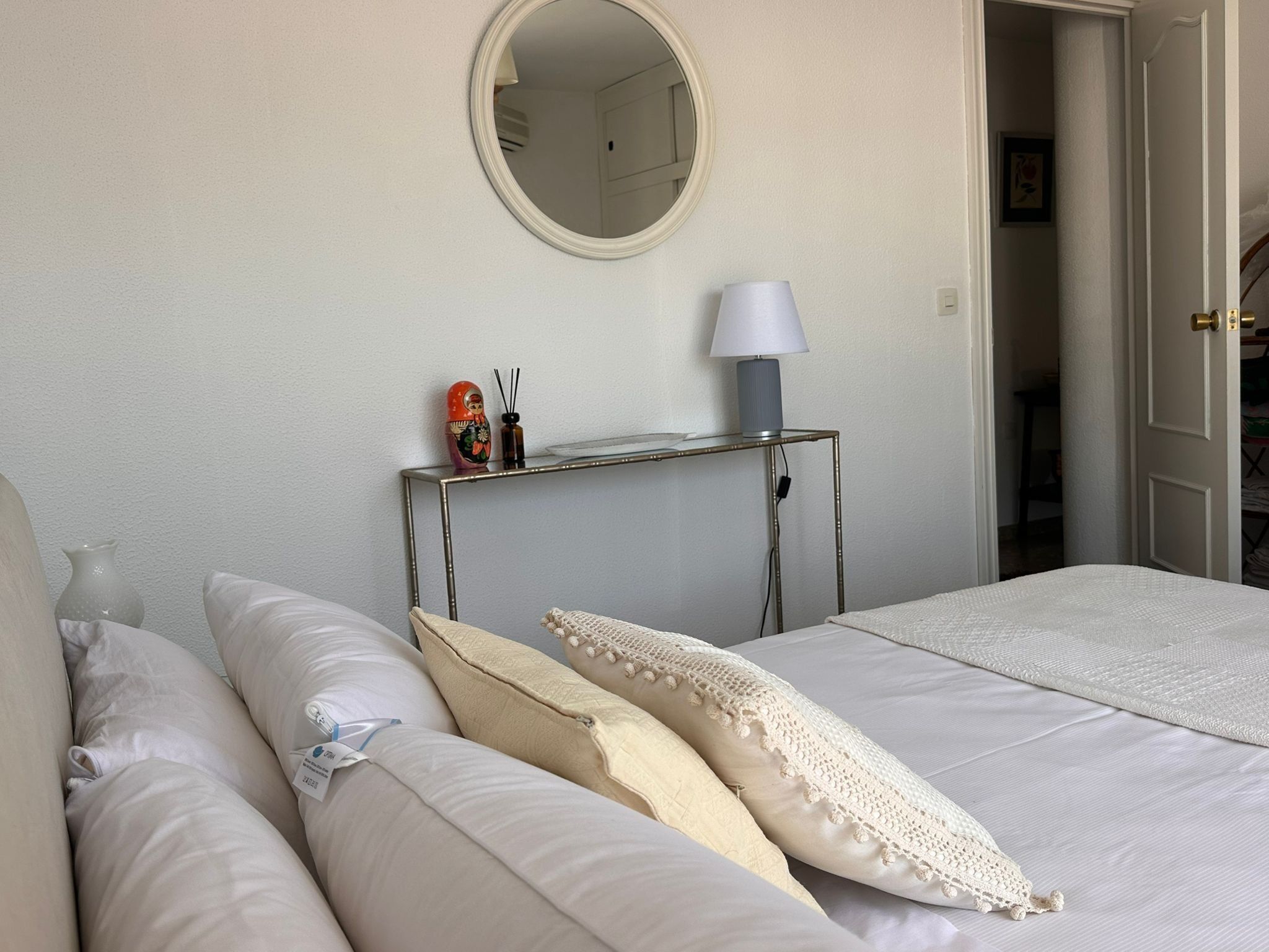 Un dormitorio con cama, almohadas, mesita de noche con lámpara, espejo y puerta que da a otra habitación.