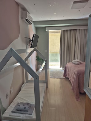 Quarto de hotel com beliche, uma escrivaninha com computador, planta em vaso, cama com cobertor rosa e vista para o exterior.