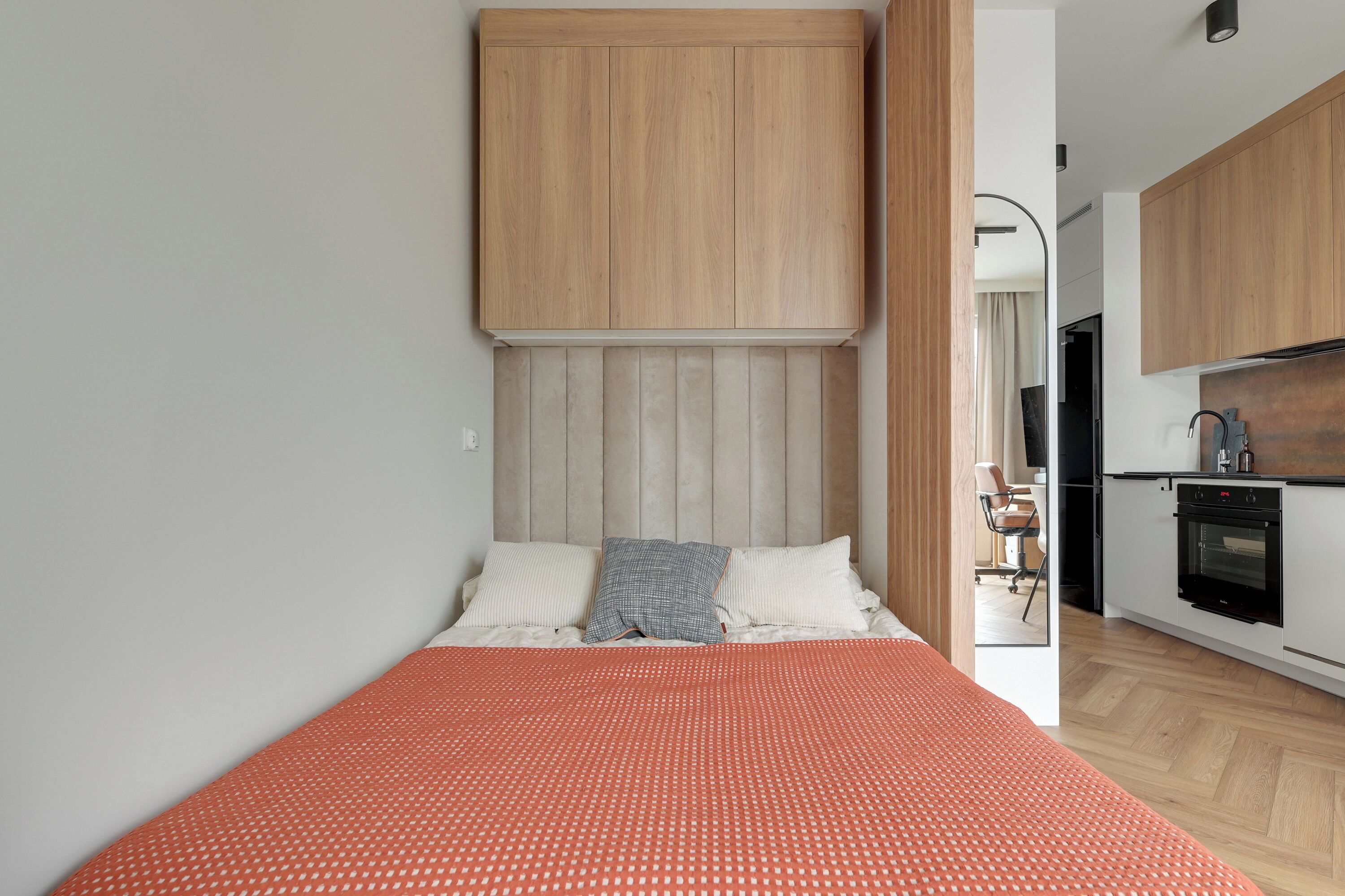 Quarto compacto com cama, cabeceira de madeira e uma kitchenette ao fundo.