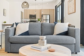 Ein modernes Wohnzimmer mit einem grauen Ecksofa, einem runden Couchtisch mit Büchern und einer Kerze, und einer Küche im Hintergrund.