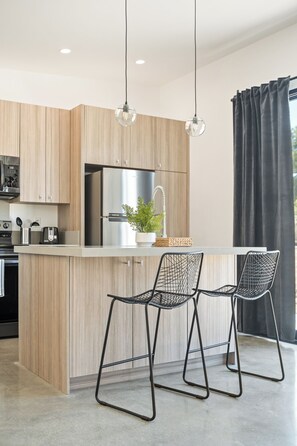 Eine moderne Küche mit Holzausstattung, Edelstahlgeräten, einer zentralen Kochinsel inklusive Barhocker, Pendelleuchten und Blick ins Freie.