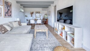 Una sala de estar con un sofá, una mesa de centro, un televisor en un estante blanco y un comedor con sillas y una mesa.