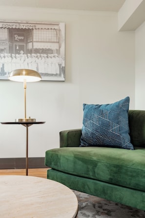 Un sofá verde con un cojín estampado, una mesa redonda de madera y una lámpara de pie con base dorada.