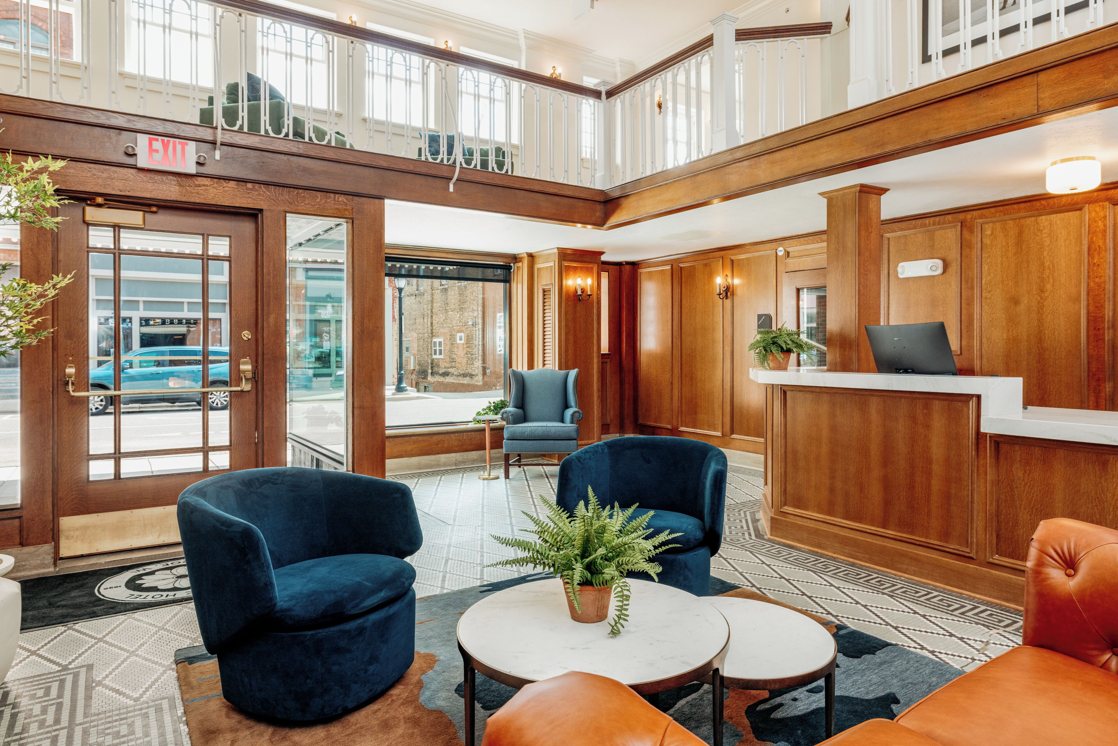 Un hall d’hôtel avec un comptoir d’accueil, un espace salon avec des fauteuils, une plante en pot et une vue sur la rue à travers les portes vitrées.