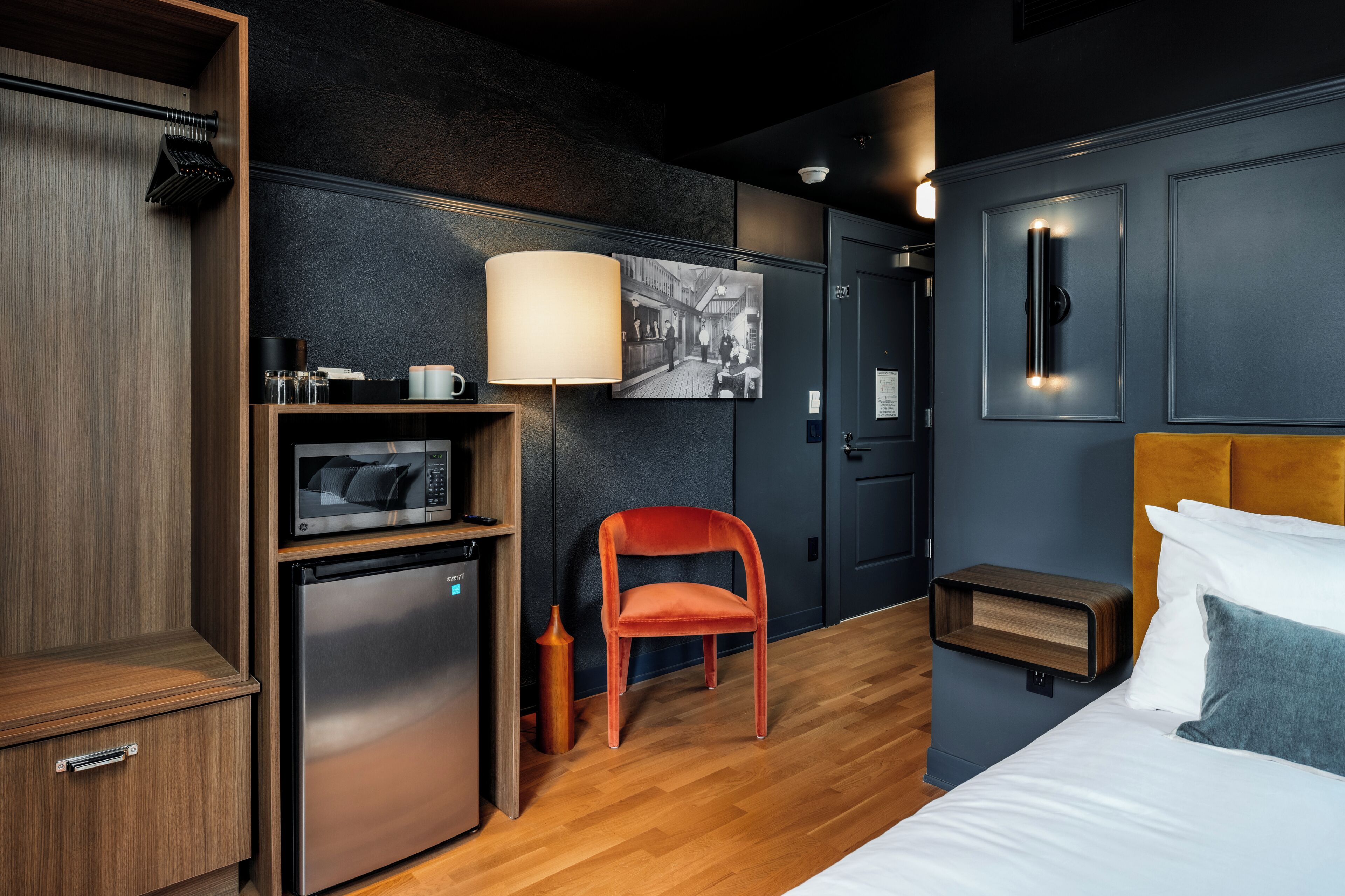 Une chambre d’hôtel compacte comprenant un lit, un bureau avec un four à micro-ondes, un réfrigérateur, une chaise, une table de chevet et une lampe fixée au mur.