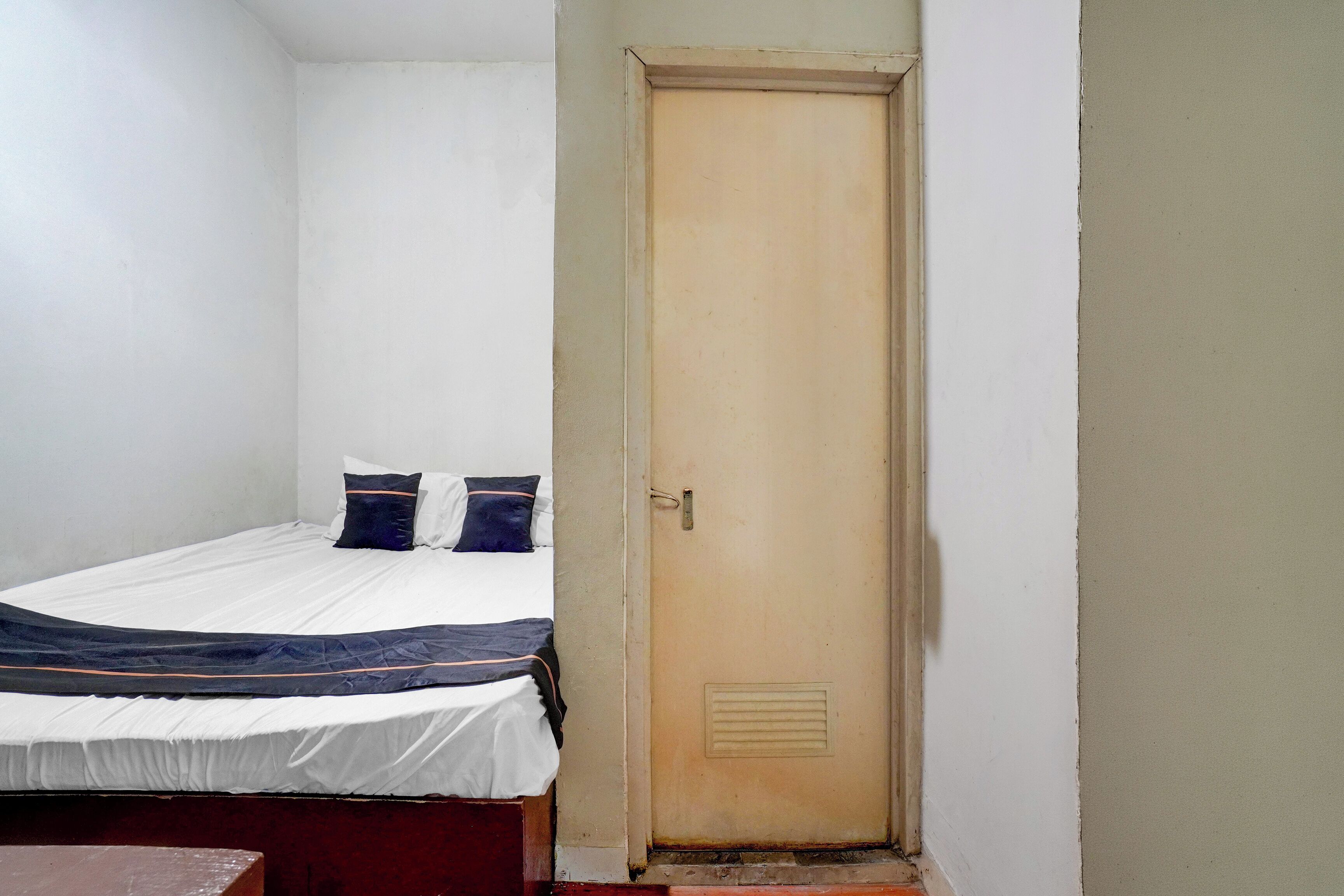 Un letto singolo con materasso bianco e biancheria da letto scura, una porta di legno e un piccolo tavolo.