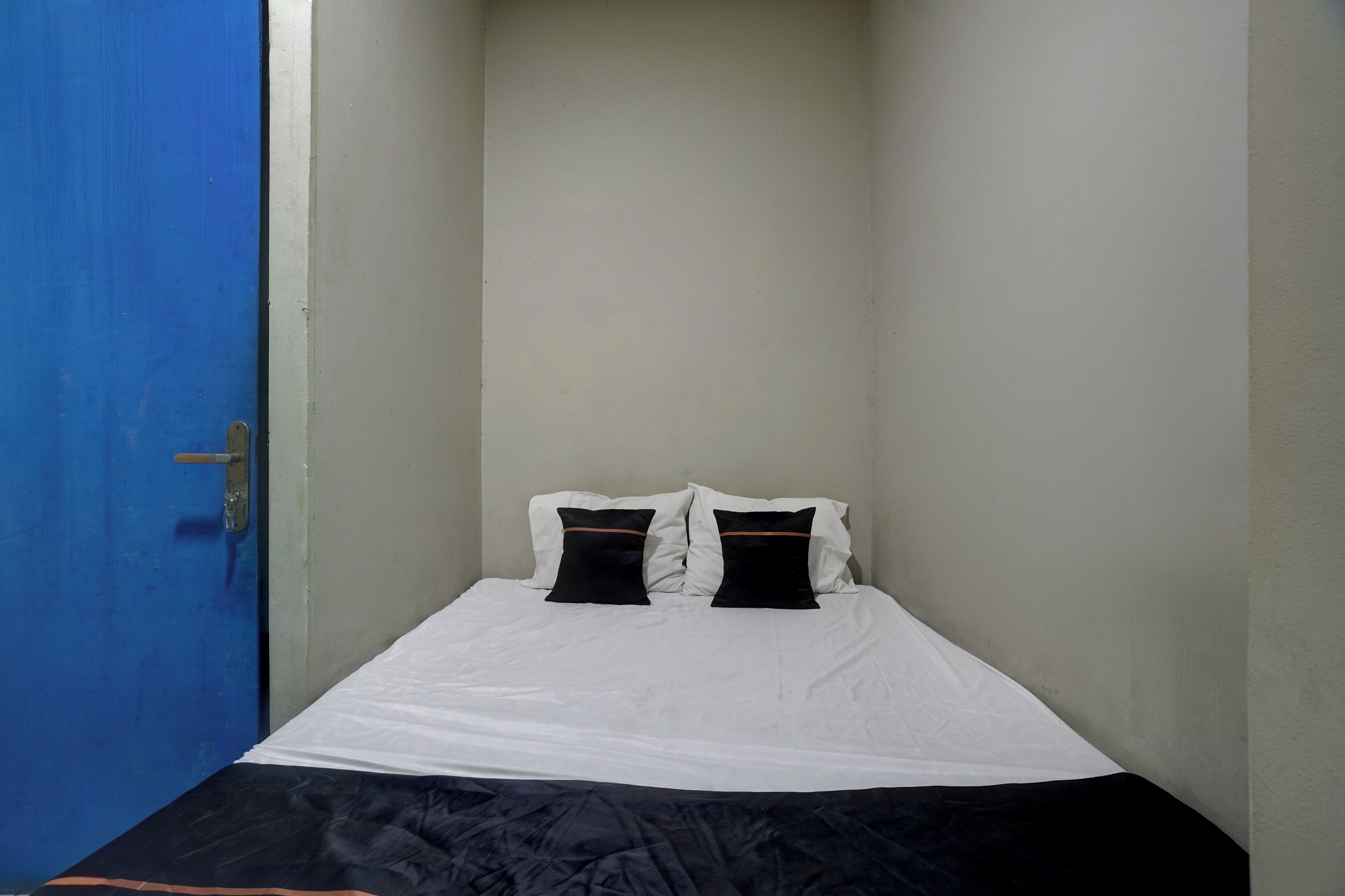 Una stanzaetta con un letto singolo, una porta blu, pareti bianche e una coperta a letto bianco e nero.