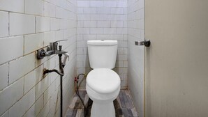 A small, tiled bathroom with a white toilet, a visible water pipe, and a door. - Hotel O near RS Husada formerly Pondokan (Jakarta)