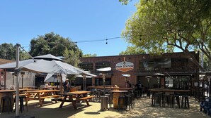 An outdoor seating area with wooden tables and benches, a brick building with a sign reading 'PARK STATION,' and string lights overhead.
