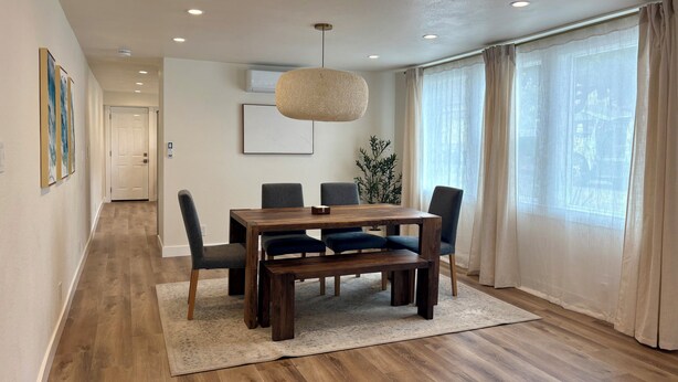 A dining area with a wooden table, chairs, a hanging light fixture, a plant, and a view through large windows.