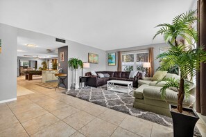 Ein Wohnzimmer mit einer braunen Ledersofa, einem weißen Couchtisch und einem grünen Sofa. Im Hintergrund ist ein Essbereich sowie eine große Topfpflanze und ein gemustertes Teppich zu sehen.