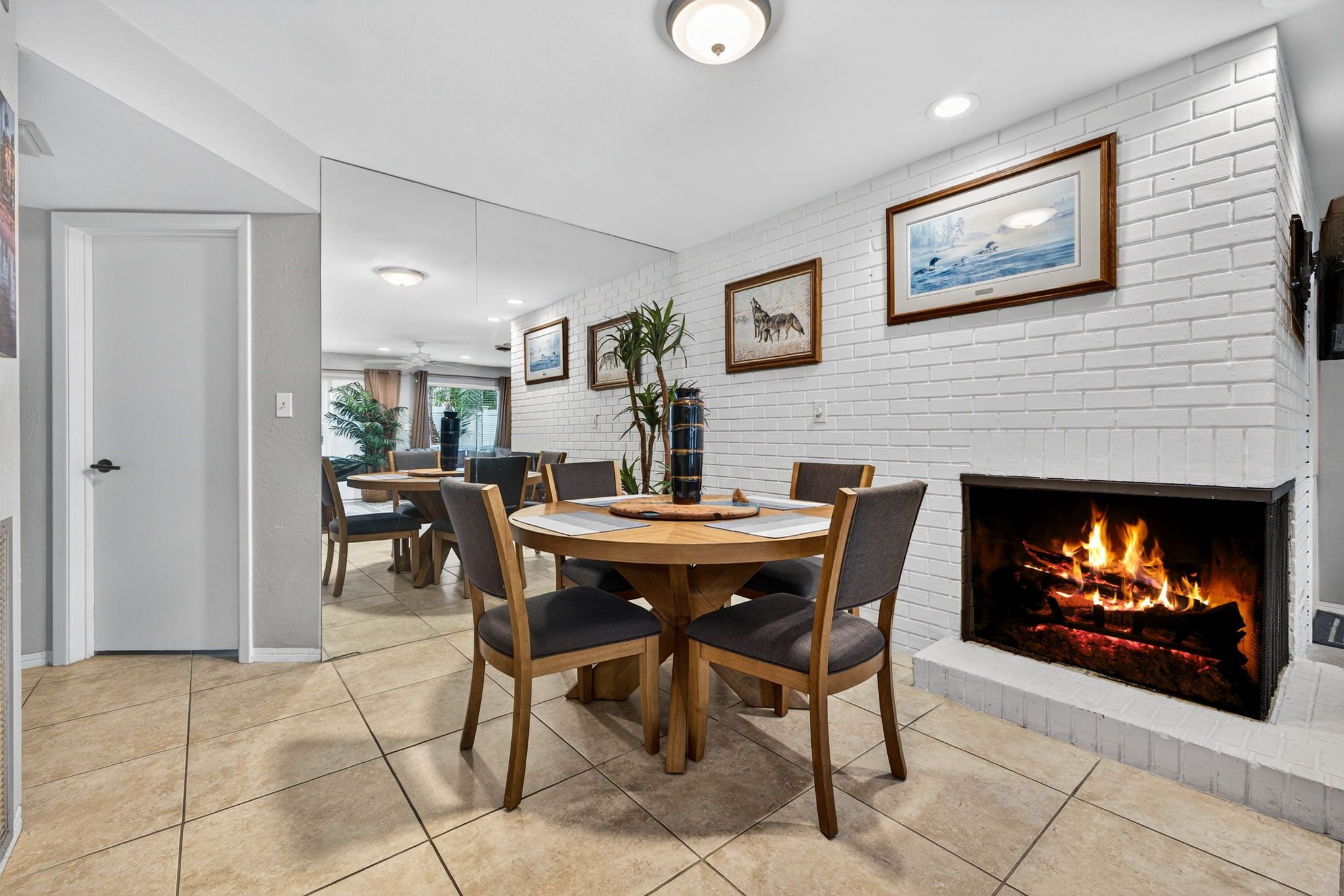 A dining area with a round wooden table and chairs, a white brick fireplace, and framed artwork on the walls.