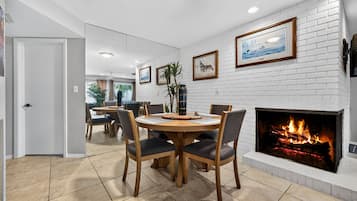 A dining area with a round wooden table and chairs, a white brick fireplace, and framed artwork on the walls.