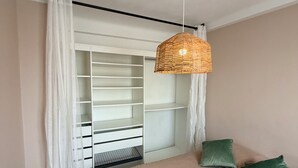 Quarto com cama, luminária de vime suspensa, armário com prateleiras e cortinas leves.