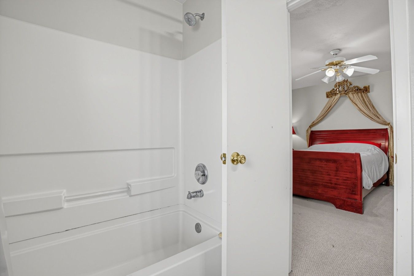 Casa de banho com banheira, chuveiro e porta que dá para um quarto com cama e ventoinha no teto.