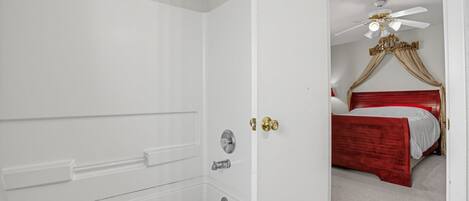 Casa de banho com banheira, chuveiro e porta que dá para um quarto com cama e ventoinha no teto.