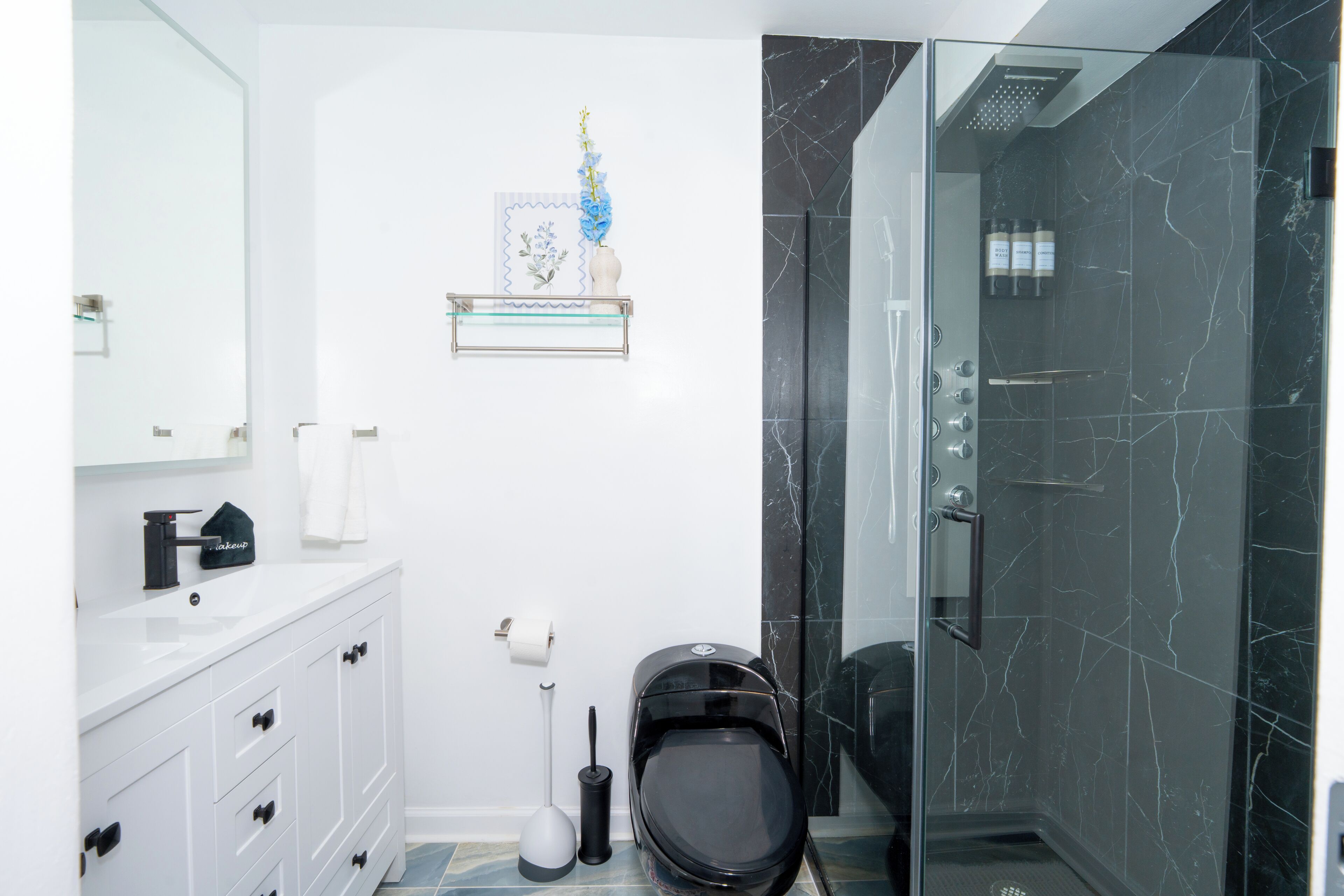 A modern bathroom with a glass-enclosed shower, white vanity with drawers, a toilet, and a small shelf with toiletries.