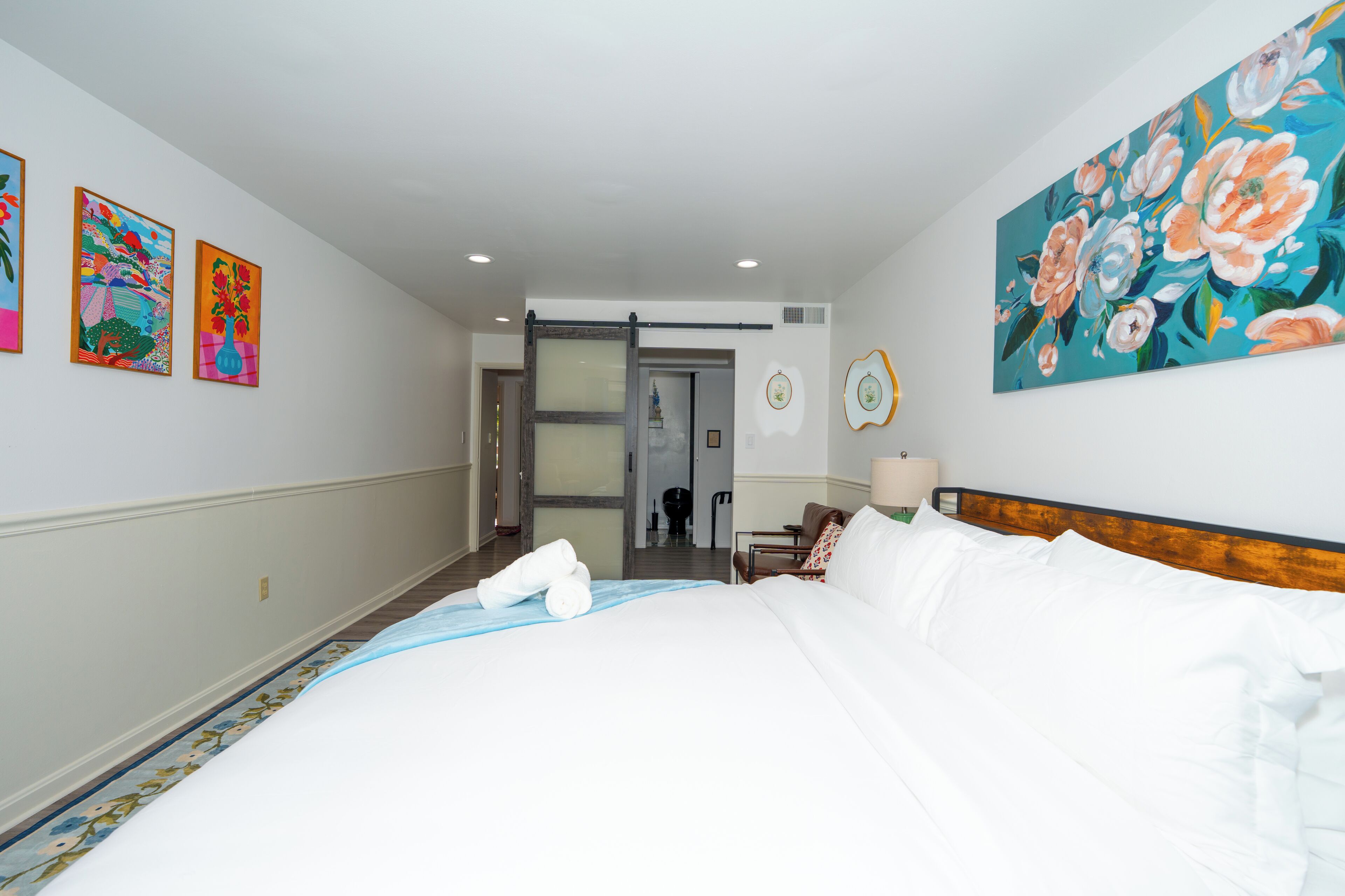 A hotel room with a bed, a door, a painting, and a wall-mounted picture frame.