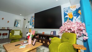 A living room with a flat-screen TV, green chairs, a wooden coffee table, and a decorative vase with flowers. - Spacious 3BR condo in Alexandria w/ 100" TV and free parking (Alexandria)