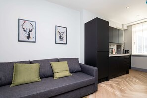 A modern living room with a grey sofa, two green cushions, and a kitchen area with dark cabinets and a window. - Edinburgh City Centre & Shore - Free Parking -WiFi (Edinburgh)