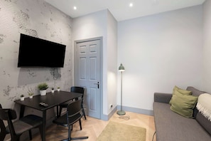 Ein Wohnzimmer mit grauem Sofa, einem schwarzen Fernseher an der Wand, einem Esstisch mit Stühlen und einer Stehlampe.