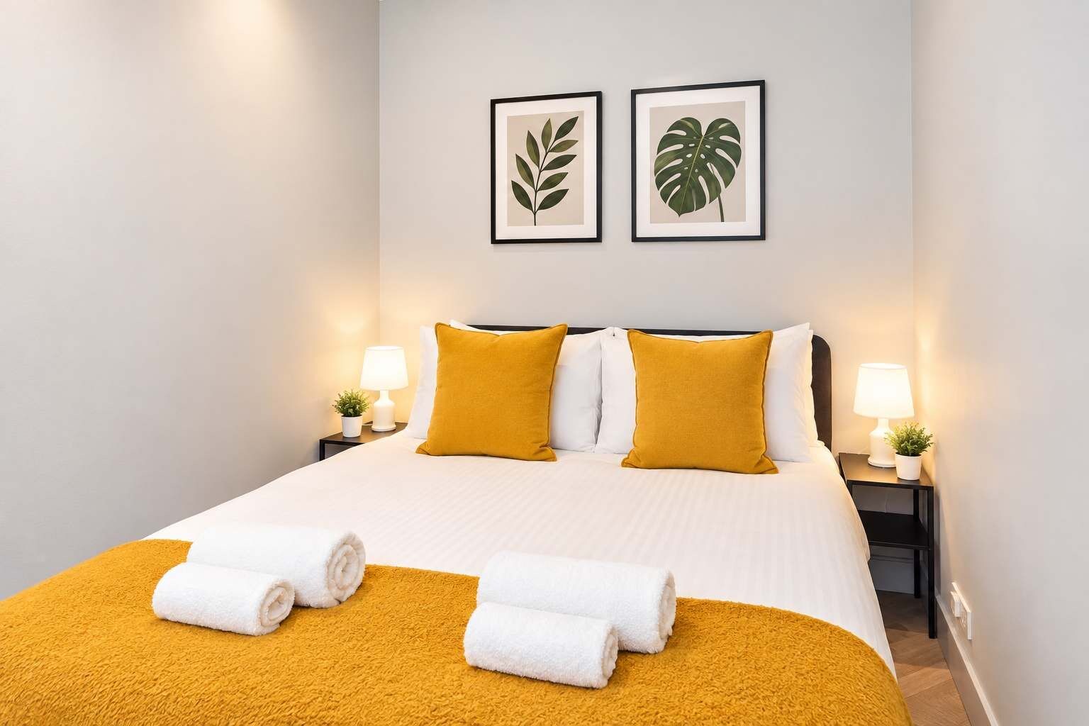 A hotel room with a bed, two pillows, two nightstands, two lamps, two framed pictures on the wall, and two towels on the bed.