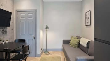 A compact living space with a dining table, chairs, a sofa, a floor lamp, and a television mounted on the wall.