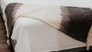 A bed with a white and dark blanket, a window with blinds, and a white wall. - The Lodge, Algonquin Woods (Perry)