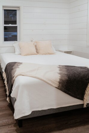 Ein Bett mit weißer und dunkler Decke, ein Fenster mit Jalousien und eine weiße Wand.