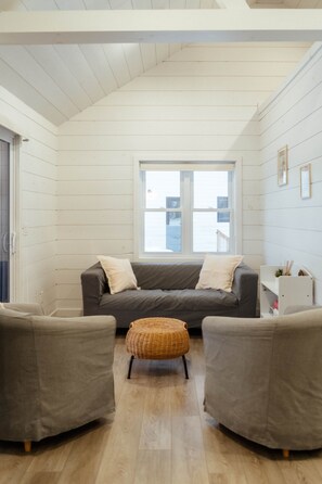 A room with a grey sofa, two armchairs, a wicker ottoman, a window with a view, and a white shelf with items. - Boreal Retreat, Algonquin Woods (Perry)