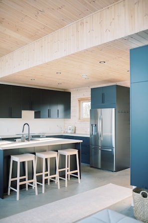 A modern kitchen with wooden ceiling, dark cabinets, a stainless steel refrigerator, and three white stools.