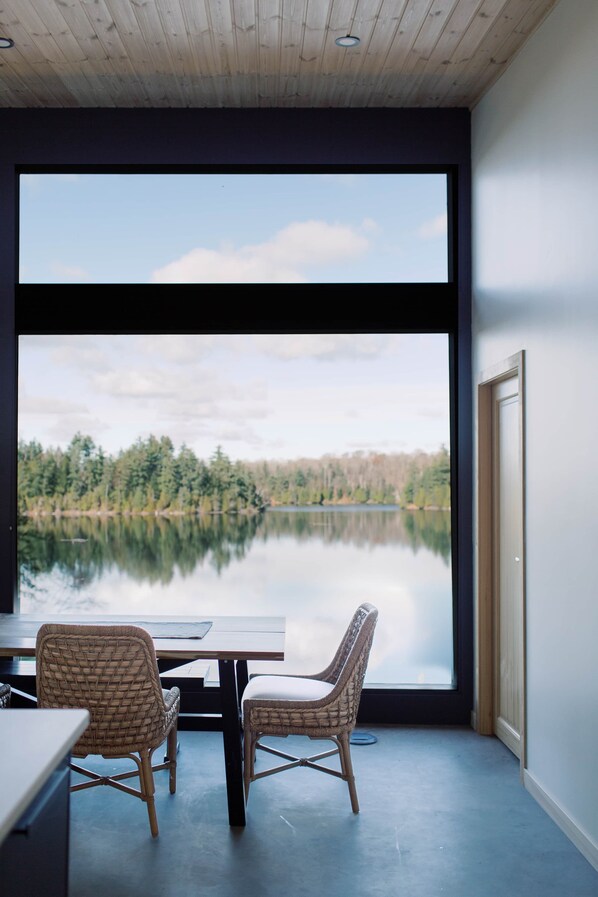 A room with a large window, two wicker chairs, a table, and a view of a lake and forest.