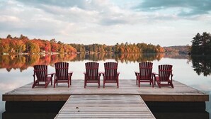 A wooden dock with five red Adirondack chairs facing a calm lake, surrounded by autumn-colored trees. - Maple Haven, Algonquin Woods (Perry)