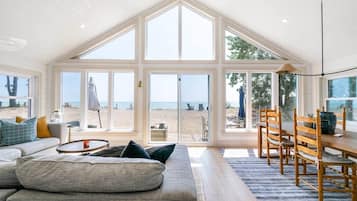 Un vaste espace de vie avec une grande fenêtre donnant sur une plage, une table à manger en bois et un coin salon confortable.