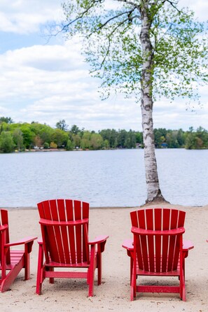 Drei rote Adirondack-Stühle an einem sandigen Strand, im Hintergrund ein See und Bäume.