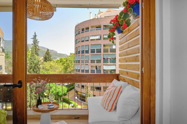 A balcony with a view of a building, a wooden railing, a white sofa, and a small table with a vase and glass. - Marbella Sky Retreat, El Nido Suite (Marbella)