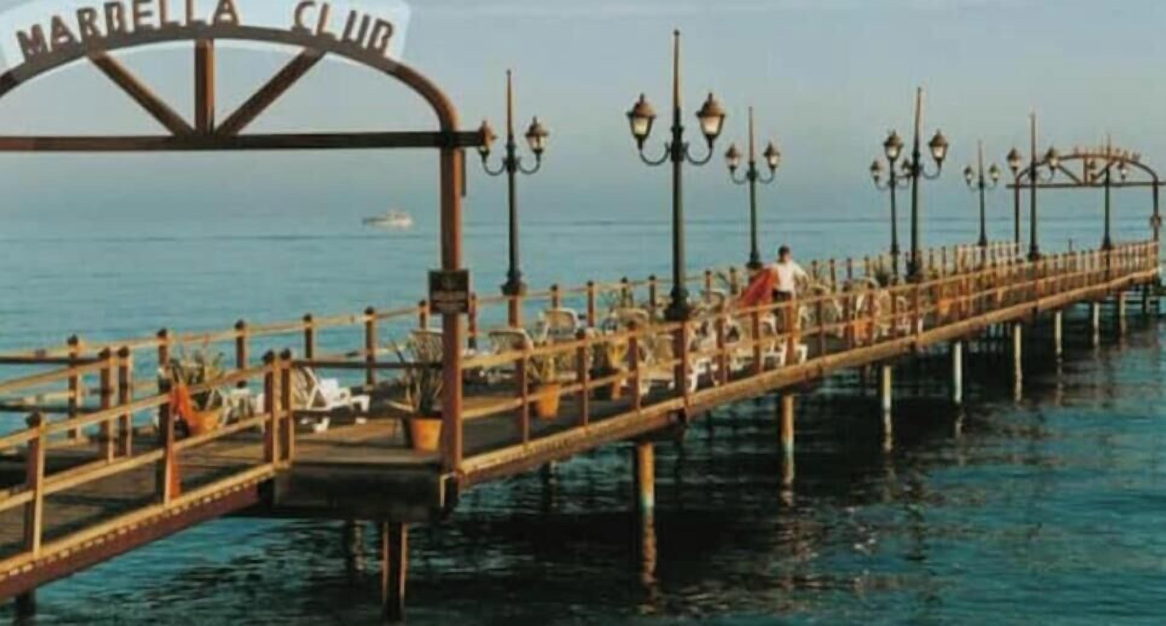 Een steiger met een bord waarop 'MARBELLA CLUB' staat, en een rij stoelen en tafels.