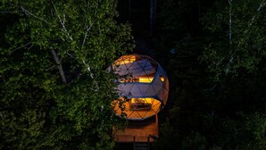 Eine geodätische Kuppel mit Holzdeck und einer darüberführenden Leiter, umgeben von dichtem Wald.