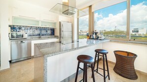 A modern kitchen with a large island, stainless steel appliances, and a view of the city through large windows. - 2 Br condo with Rooftop Pool - Doca 303 (Playa del Carmen)