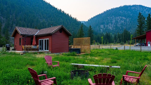 A red cabin with a patio, surrounded by green grass and mountains. - The Farmhouse at Deadman Acres (Thompson-Nicola)