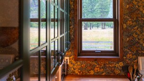 A room with a stone wall, a window with a view of trees, a wooden floor, and a kitchen area with a sink and utensils. - The Farmhouse at Deadman Acres (Thompson-Nicola)