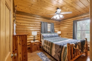 Un dormitorio tipo cabaña de madera con cama, mesitas de noche, ventilador de techo y ventana.