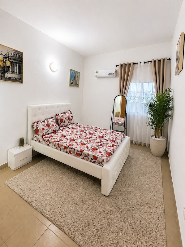 Une chambre à coucher avec un lit, une table de chevet, une plante en pot, un radiateur et une fenêtre avec des rideaux.